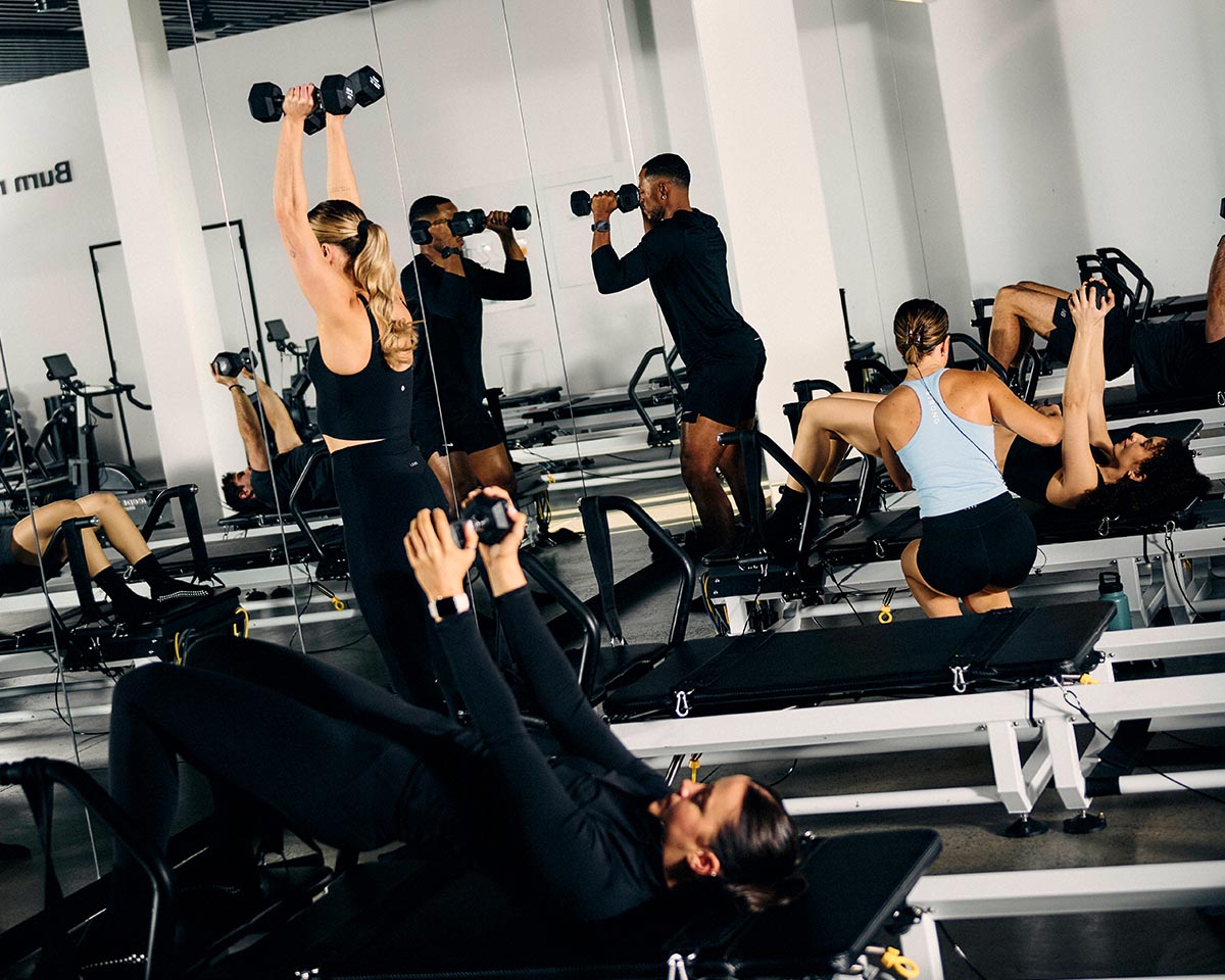 Members pressing dumbbells overhead in a full class